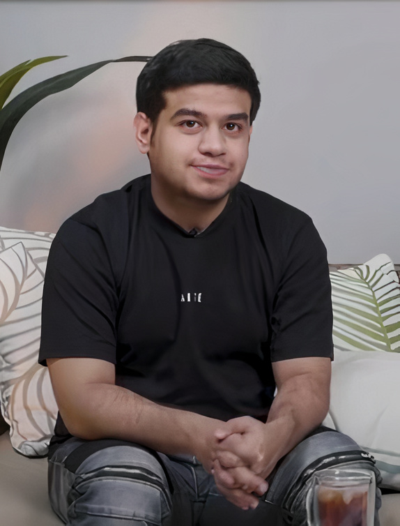 Pria muda duduk santai di sofa dengan latar belakang tanaman hijau.